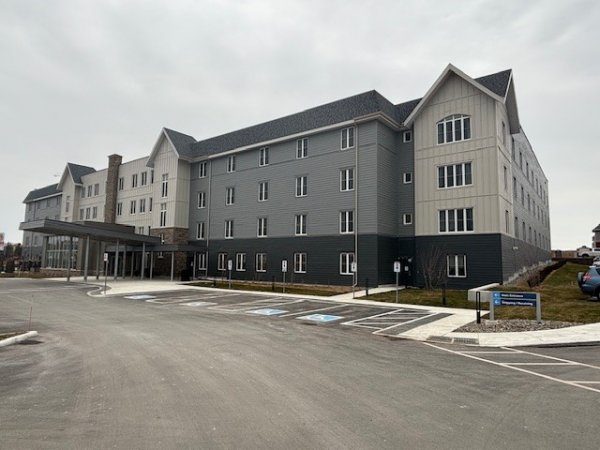 A four-story modern hotel building with gray siding, an entrance canopy, and an adjacent parking lot with several accessible parking spaces.