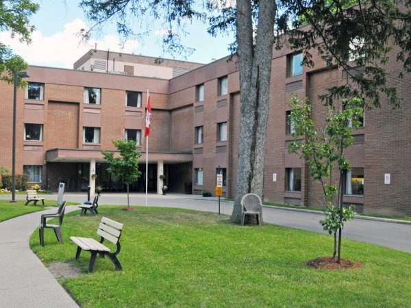 A three-story brick building with a Canadian flag at the entrance, surrounded by green lawns, benches, and trees.