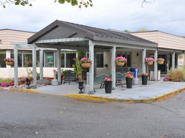 Single-story building entrance with a covered patio, benches, and hanging flower baskets. The area is surrounded by pavement and landscaped plants.