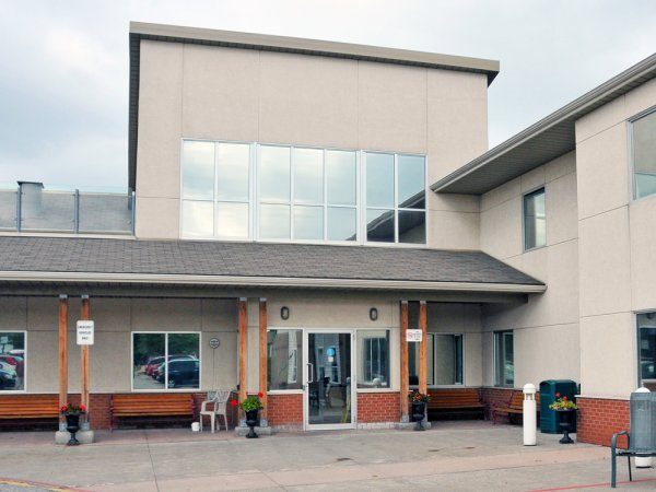 A modern, two-story building with large windows, a covered entrance, benches, flower pots, and signs near the doorway.
