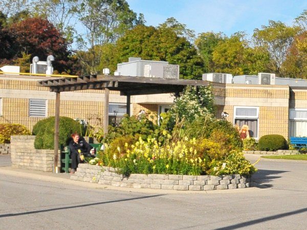 A woman sits on a bench under a wooden pergola beside a circular flower bed in front of a brick building with several windows.