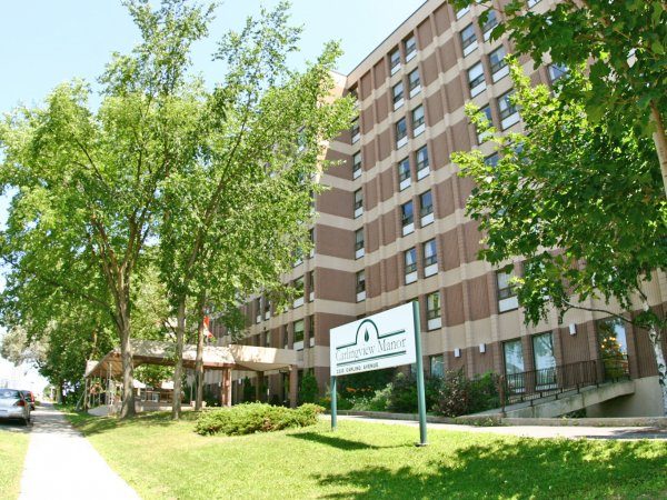 A multi-story brick building with large windows, surrounded by trees and grass, with a sign reading "Lamplight Manor" at the entrance.