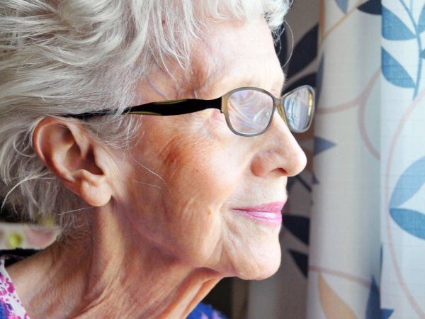 Elderly woman with glasses and white hair looks out of a window, daylight illuminating her face and patterned curtains visible in the background.