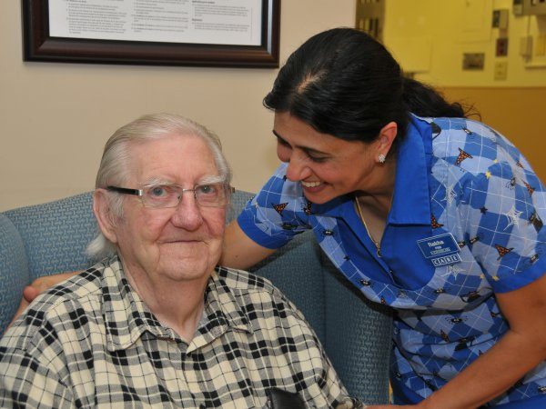 An elderly man sits in a chair while a smiling female caregiver in a blue uniform stands beside him, leaning in supportively.