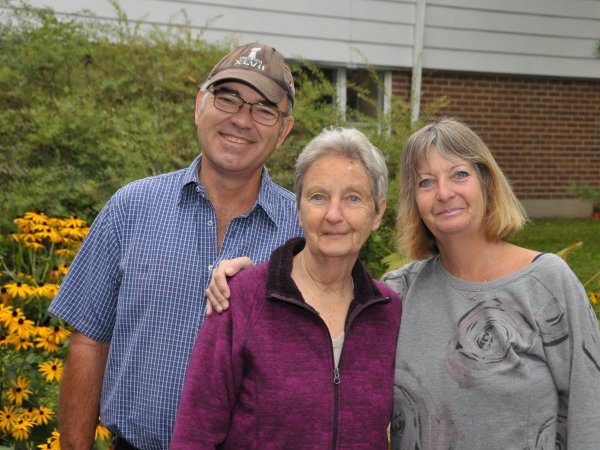 Three adults stand close together outside in front of yellow flowers and greenery, with a brick building in the background.