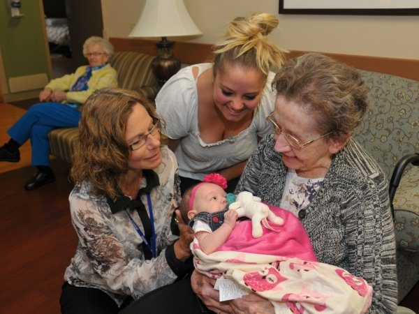 An elderly woman holds a baby wrapped in a pink blanket, surrounded by two women smiling, while another elderly woman sits in the background.
