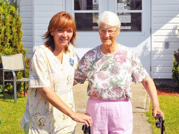 A caregiver stands beside an elderly woman using a walker outside a building on a sunny day.