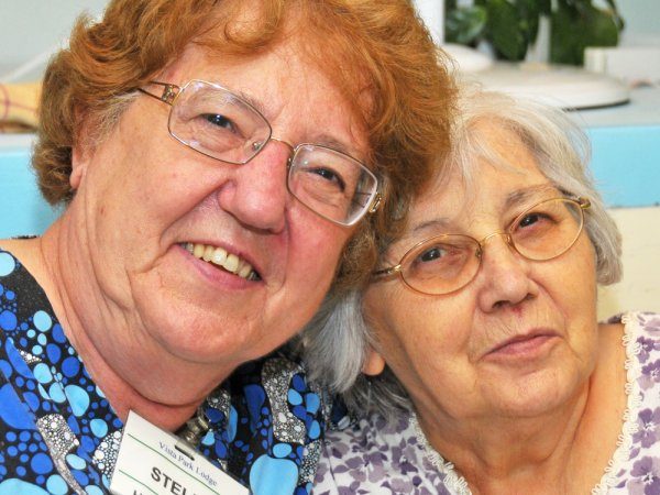 Two elderly women with glasses sit closely together, smiling at the camera. One wears a blue patterned top with a name tag, the other wears a purple and white blouse.