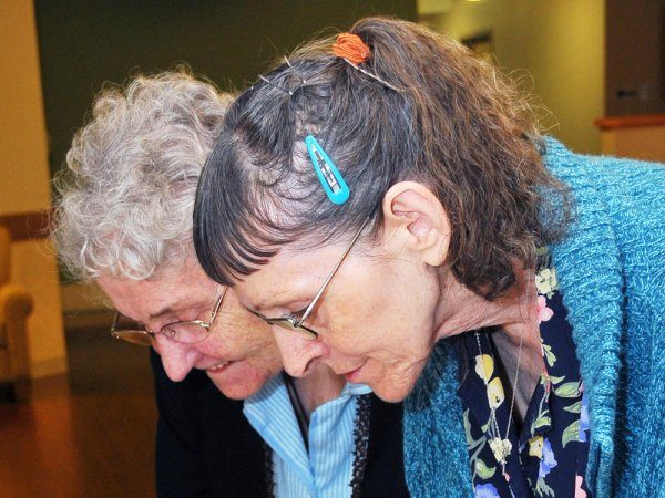 Two elderly women with gray hair, one wearing glasses and a blue hair clip, lean in closely together, looking down at something out of view.