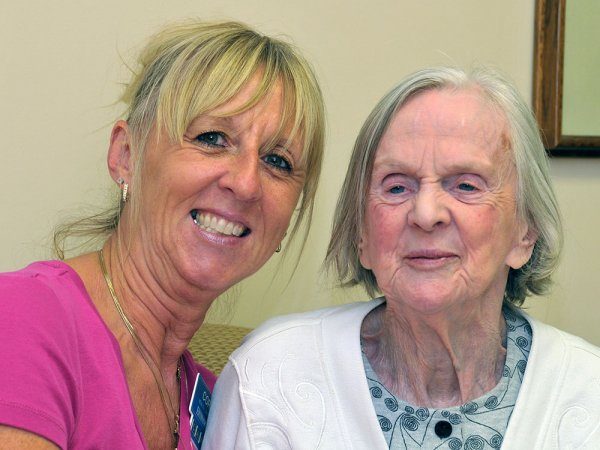 Two women, one younger and one elderly, sit closely together and smile at the camera in a well-lit indoor setting.