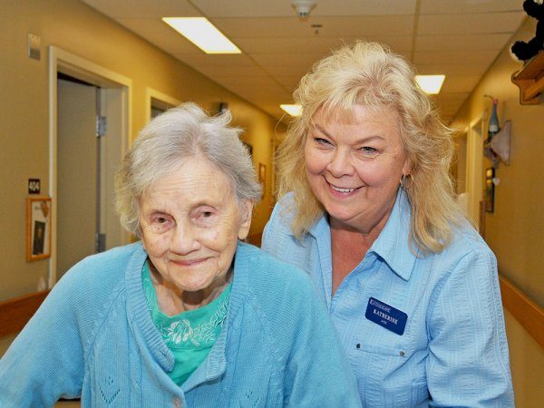 An elderly woman with gray hair stands in a hallway with a smiling female caregiver wearing a name tag. Both women are dressed in blue.