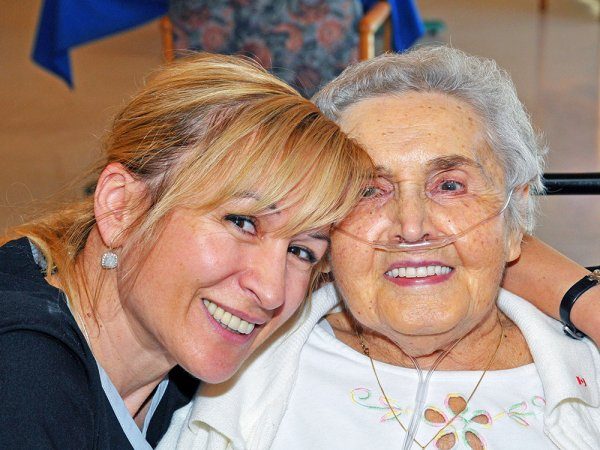 A younger woman hugs an older woman using an oxygen tube, both smiling at the camera in a brightly lit indoor setting.