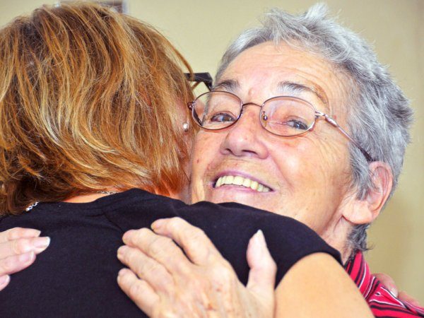 An older woman with glasses smiles while hugging another person, whose back is facing the camera.