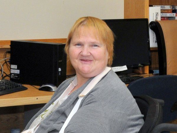 A woman with short blonde hair sits smiling in front of a computer in an office or library setting.