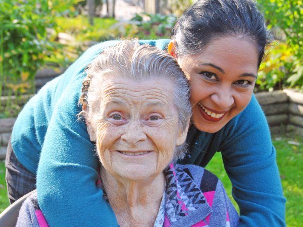 An elderly woman sits smiling in a garden while a younger woman stands behind her, hugging her shoulders and smiling at the camera.