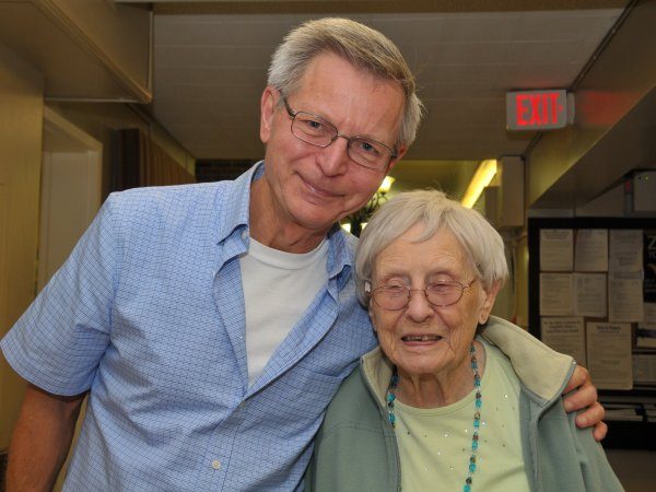 An older man and woman stand side by side indoors, smiling at the camera. The man has his arm around the woman. A red exit sign is visible in the background.