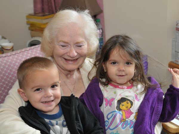 An elderly woman sits smiling with a young boy and girl on her lap. The girl is wearing a Dora shirt and holding her hair. All three are facing the camera.