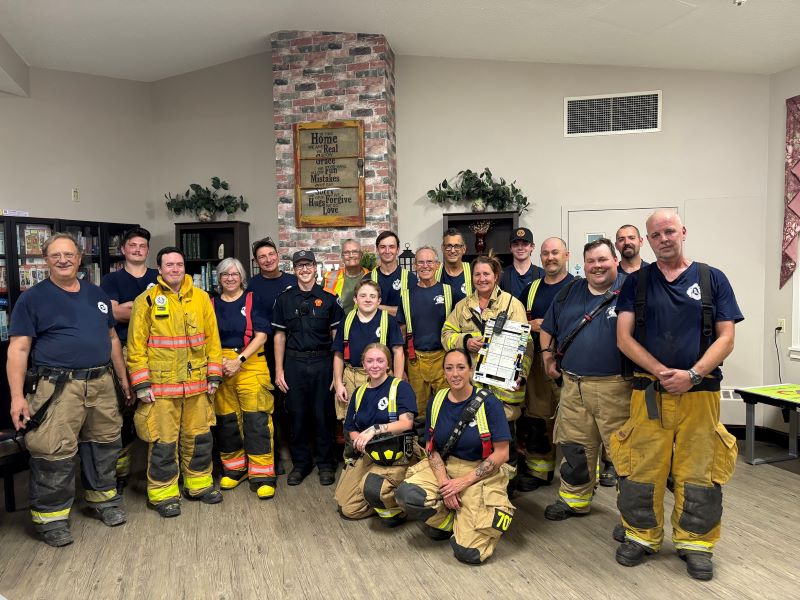 A group of firefighters and emergency personnel pose together indoors, some in full gear and others in uniforms, smiling at the camera.