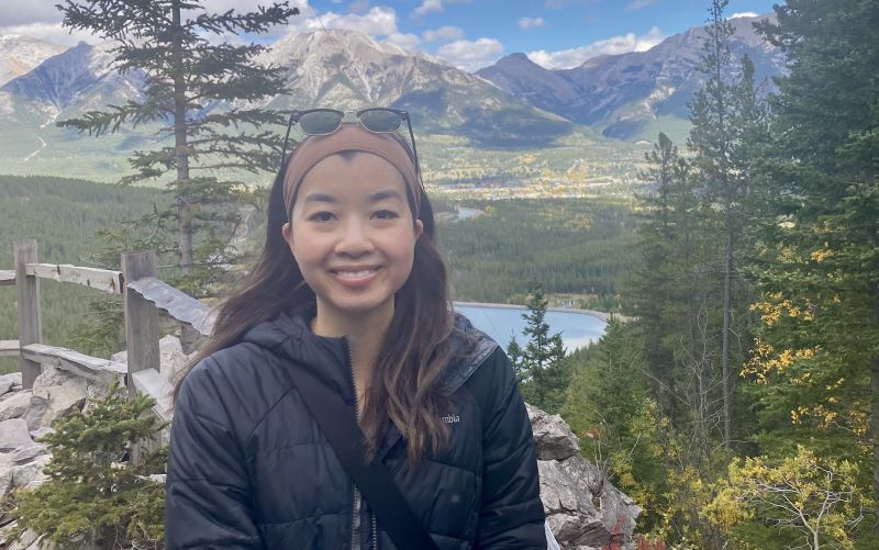 A woman wearing a black jacket and sunglasses on her head smiles in front of a mountain landscape with trees, a wooden railing, and a lake in the background.