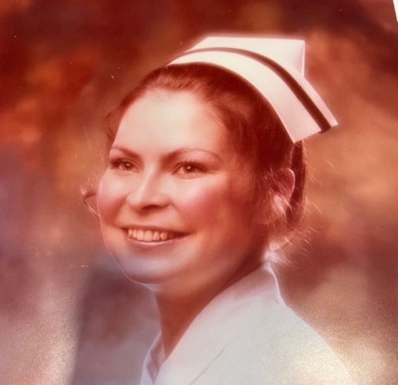 A woman wearing a white nurse uniform and cap smiles at the camera against a softly blurred background.
