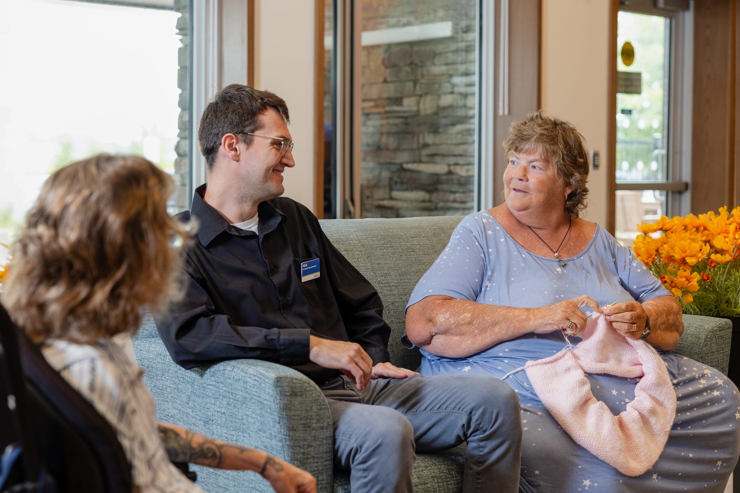 Three people sit on a couch in a bright room; one woman knits while talking to a man with a name badge, and another person listens.