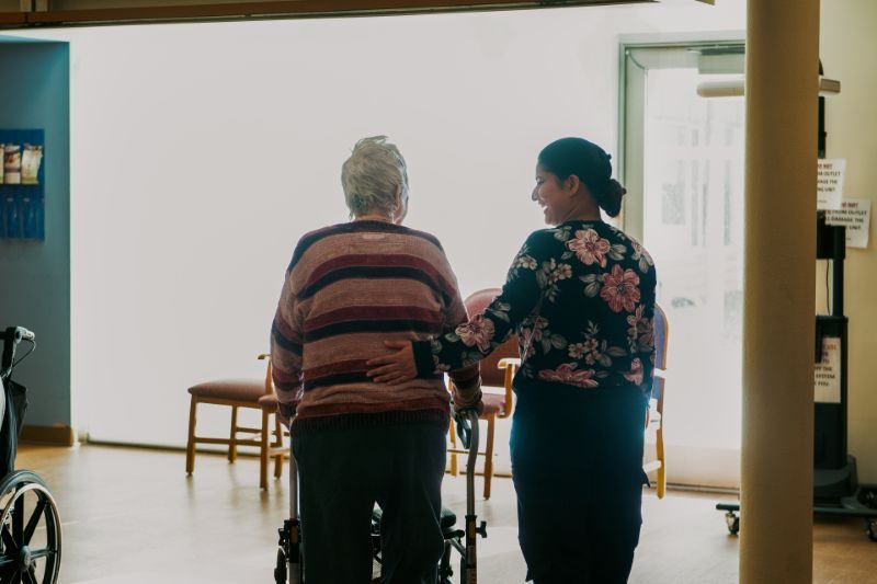 An elderly person with a walker is assisted by a caregiver as they walk through a well-lit room with chairs near the far wall.