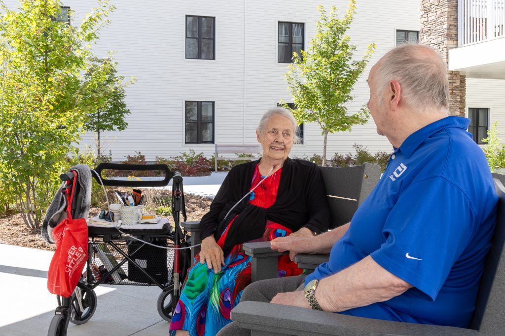 An elderly woman with an oxygen tube sits outdoors talking with an elderly man. A walker with a red bag is beside her. They are seated near a building with trees in the background.