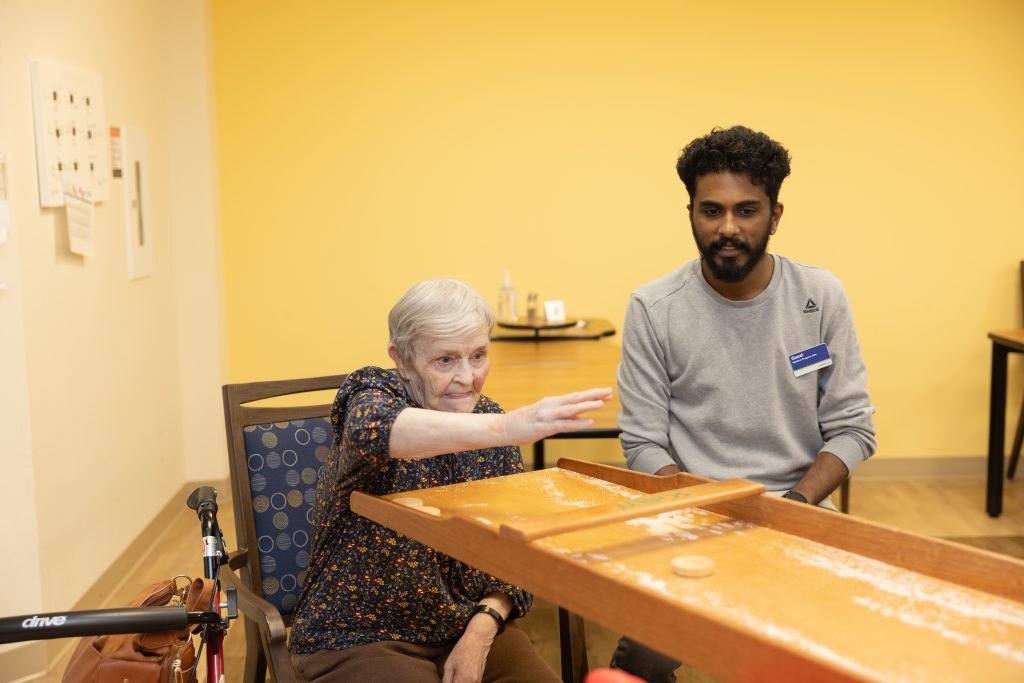 An elderly woman plays a tabletop game while a younger man observes in a brightly lit room with yellow walls.