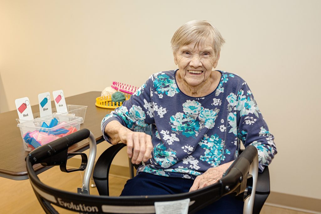 Elderly woman sitting in a walker chair, smiling at the camera, with color-coded pill containers and daily medication organizers on a table beside her.