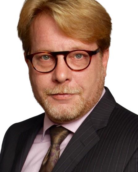 A man with light brown hair, glasses, and a trimmed beard, wearing a suit, pink shirt, and patterned tie, against a white background.