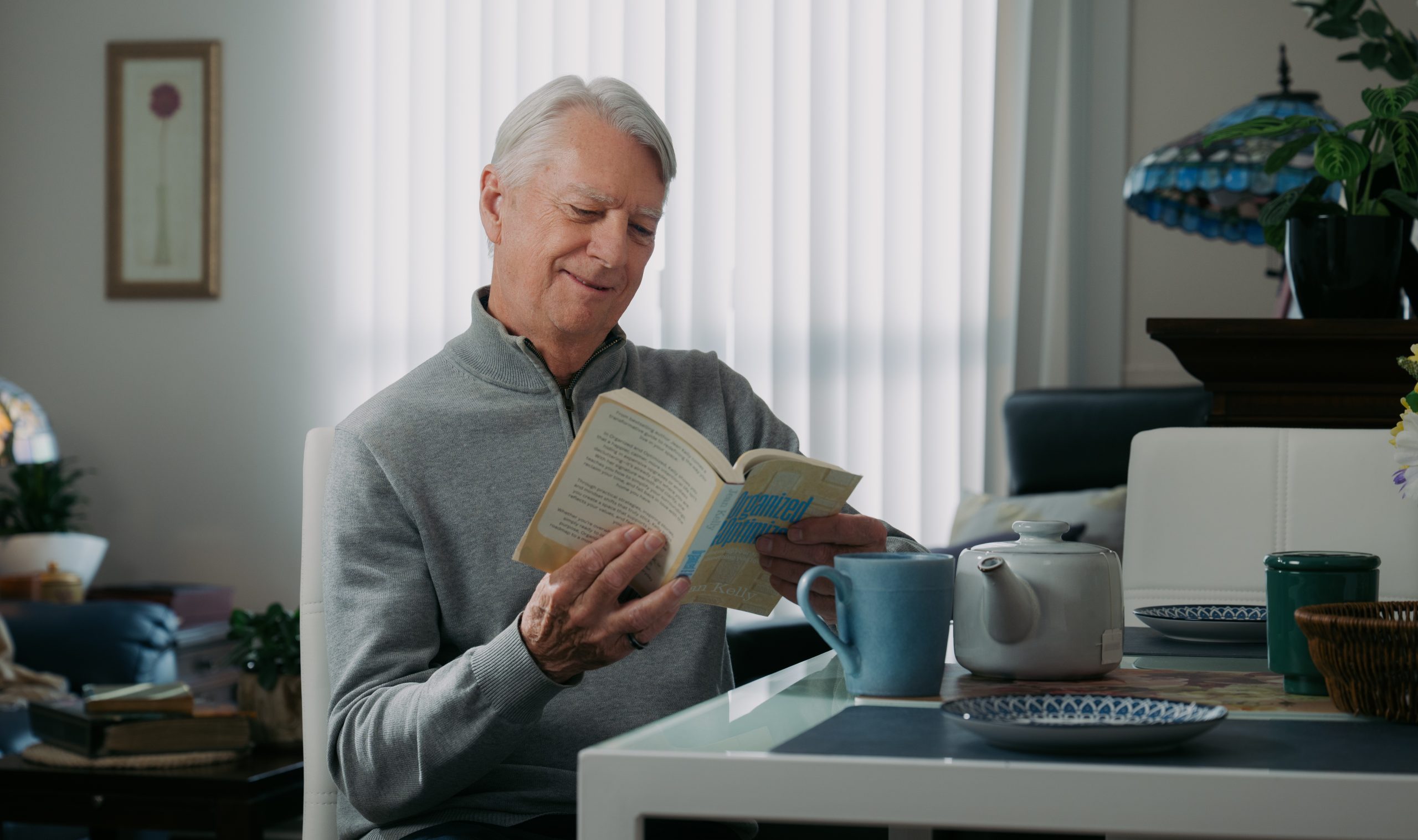 An older man with gray hair sits at a table reading a book, with a teapot, mug, and plate in front of him in a well-lit room.