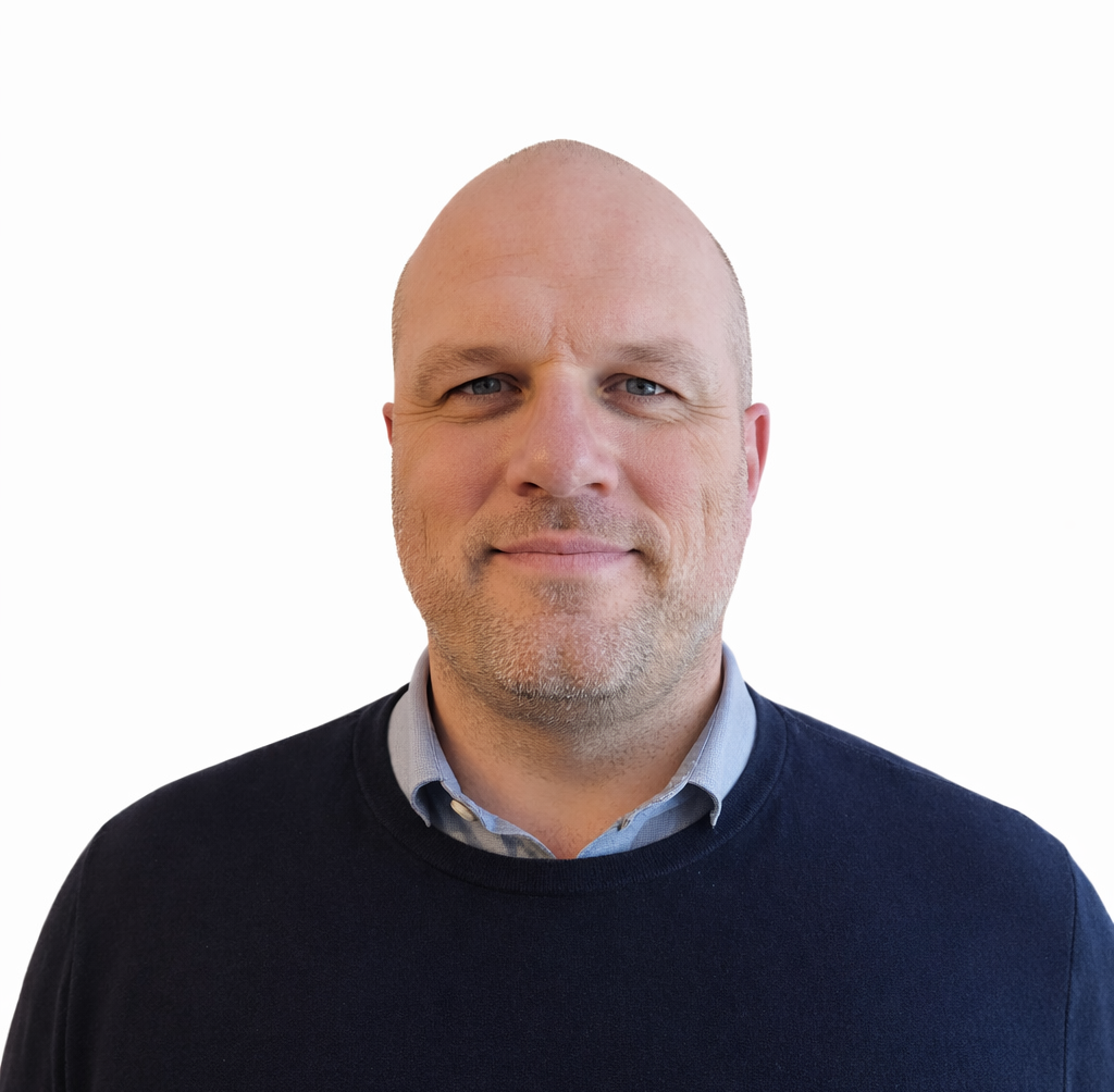 A middle-aged man with a shaved head, wearing a blue sweater over a light blue collared shirt, stands facing the camera against a plain white background.
