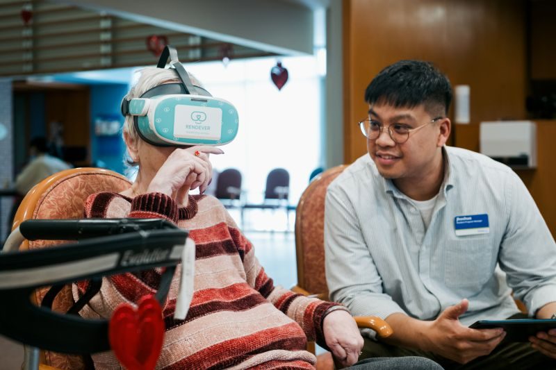 An older adult wearing a VR headset sits next to a man holding a tablet in a community space decorated with heart ornaments.