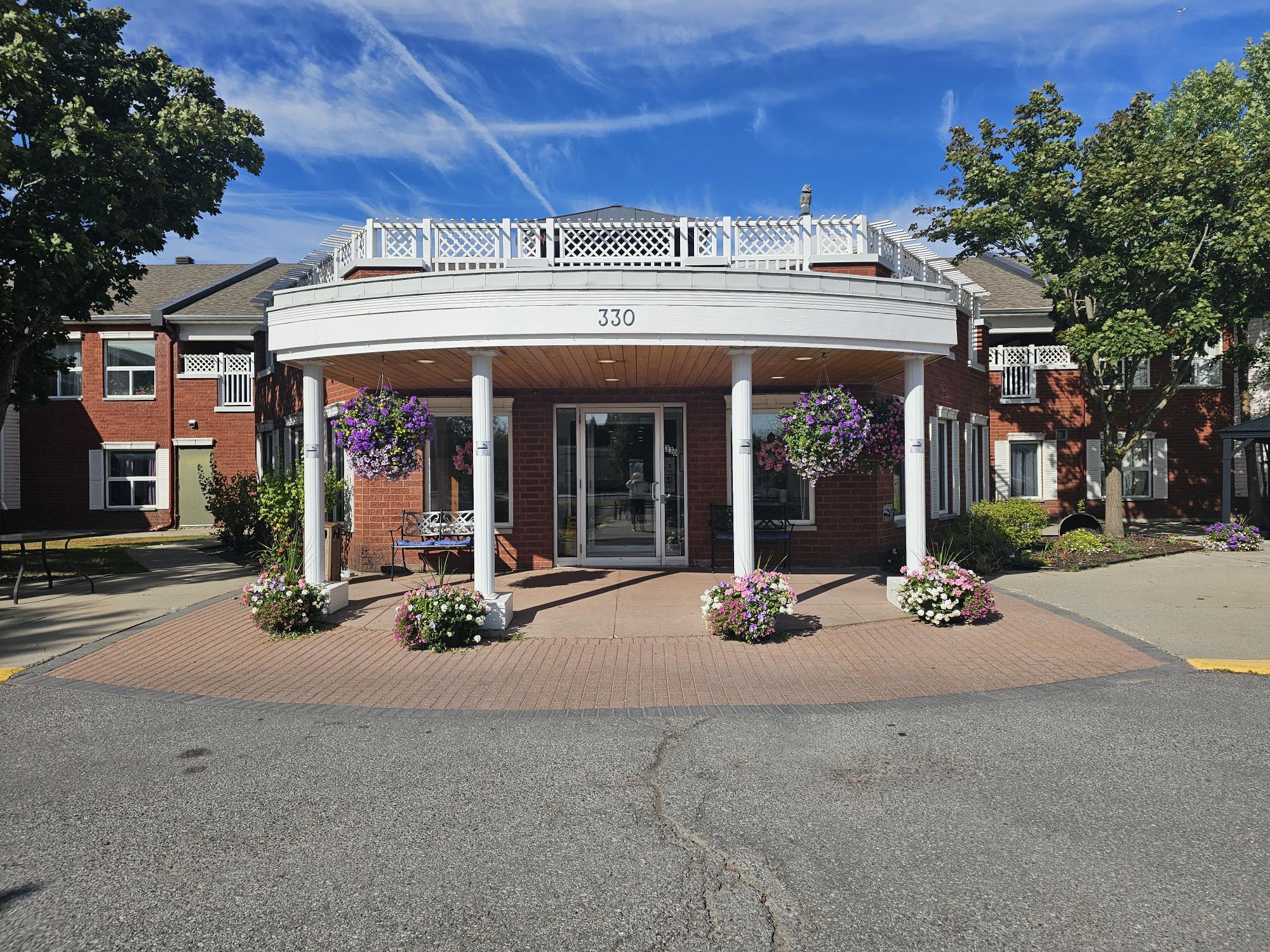A brick building with a white circular entrance, potted flowers, benches, and the number 330 above the doorway. Blue sky and trees are visible in the background.
