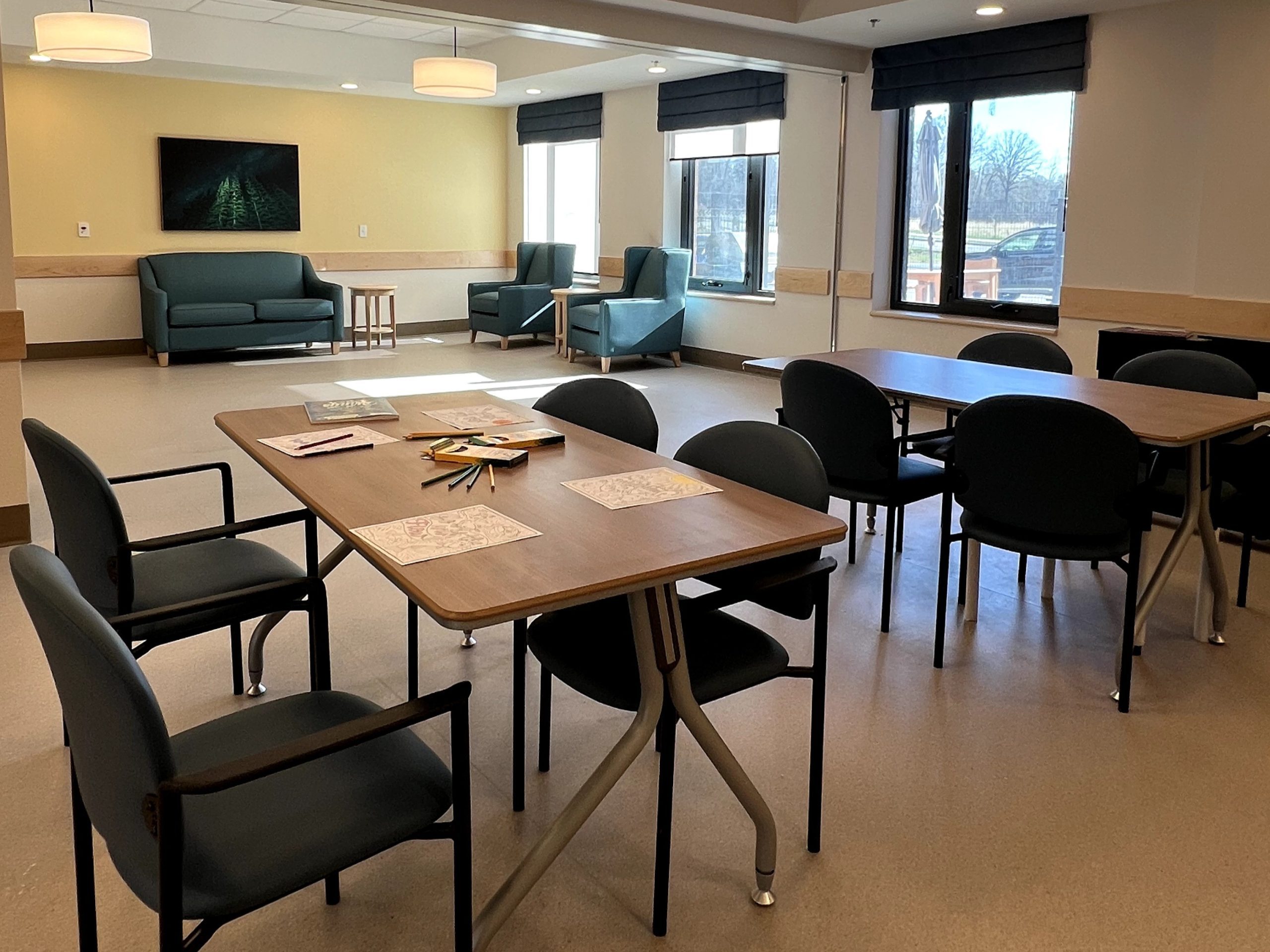 A brightly lit common room at Extendicare Beauclaire features tables, chairs, lounge seating, large windows, and papers with colored pencils on the tables.