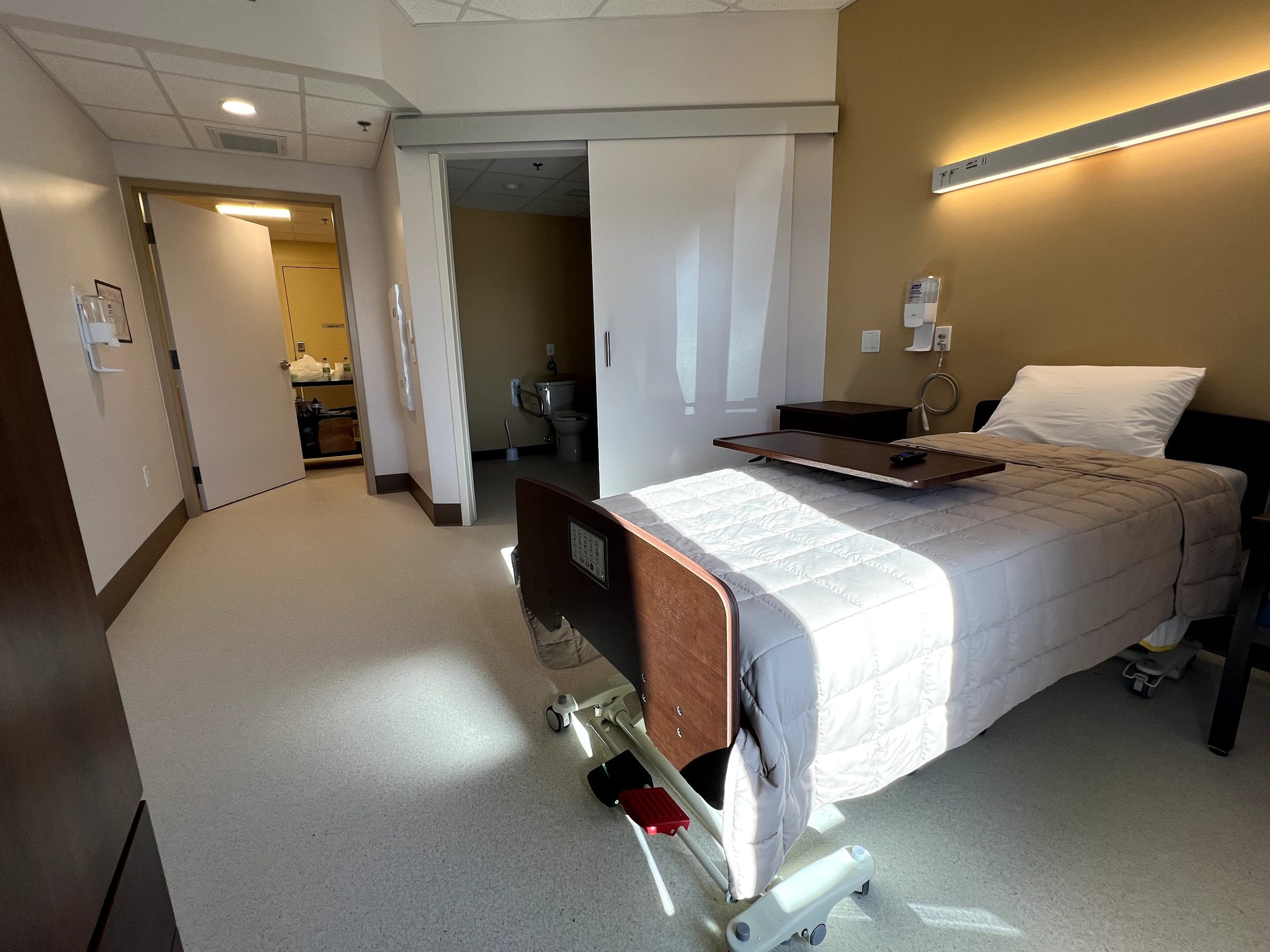 A hospital room at Extendicare Beauclaire features a single bed, light brown walls, a tray table, a pillow, and a partially open door leading to a bathroom.