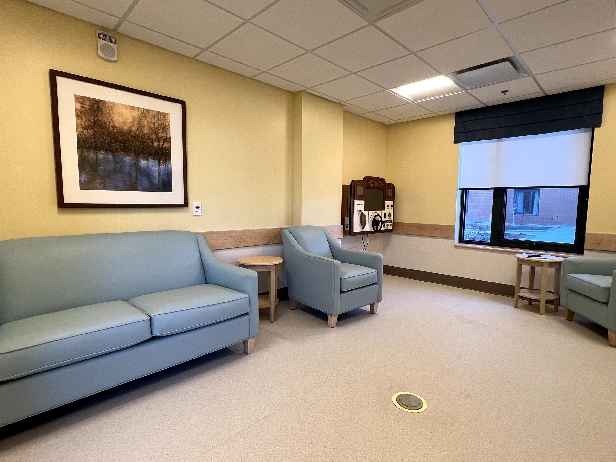 A hospital waiting room at Extendicare Beauclaire features light blue seating, a framed landscape photo on the wall, a window with a dark valance, and medical equipment on the far wall.