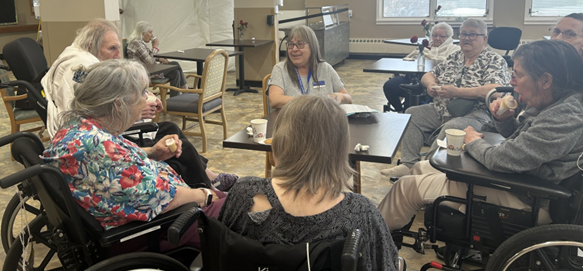 A group of older adults, some in wheelchairs, sit around tables in a common room, eating and talking together.