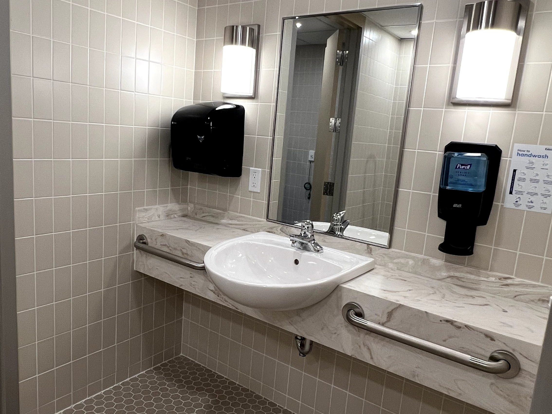 Accessible public restroom sink area at Extendicare Beauclaire with a mirror, soap dispenser, paper towel dispenser, grab bars, and tiled walls and floor.
