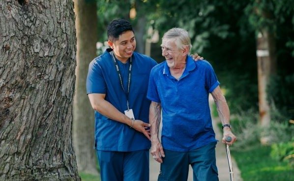A healthcare worker assists an older man with a cane as they walk outdoors on a sidewalk lined with trees.
