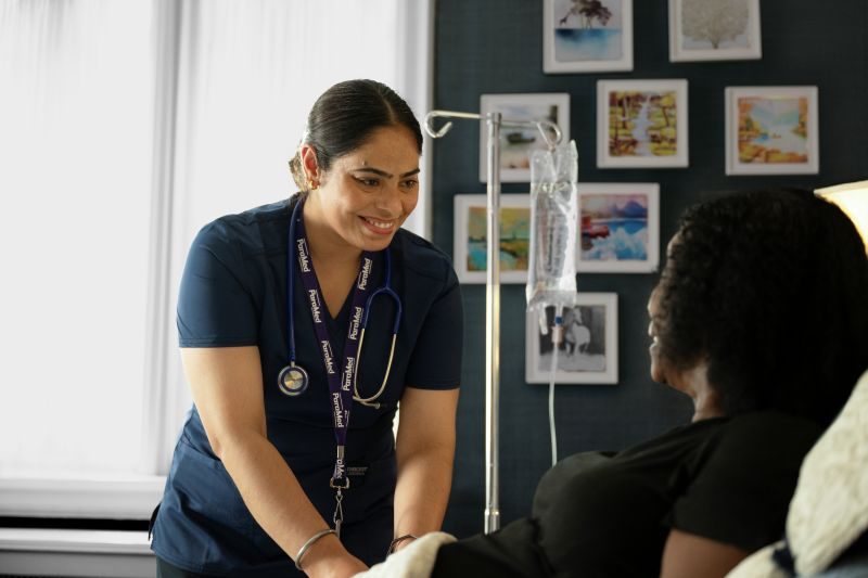 A nurse in blue scrubs smiles while attending to a patient lying in bed with an IV stand nearby; framed photos hang on the wall in the background.