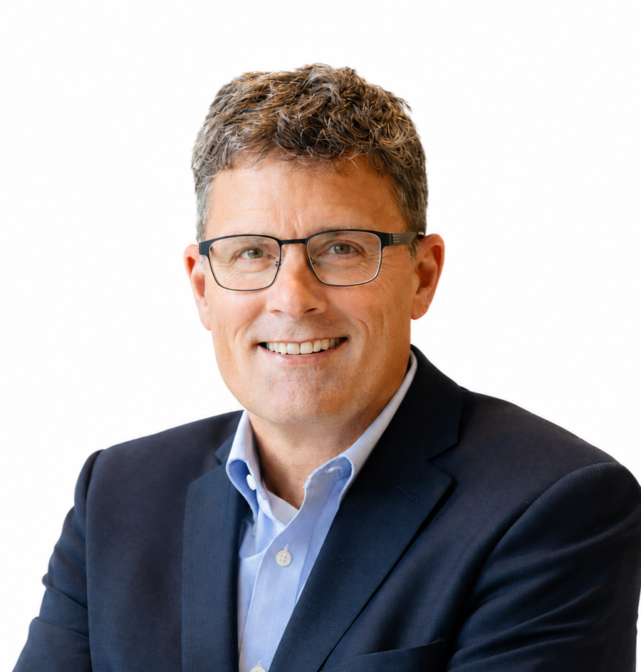 A man with short, curly brown hair and glasses, part of the Extendicare leadership team, is wearing a navy blue suit jacket over a light blue shirt, smiling at the camera against a plain white background.