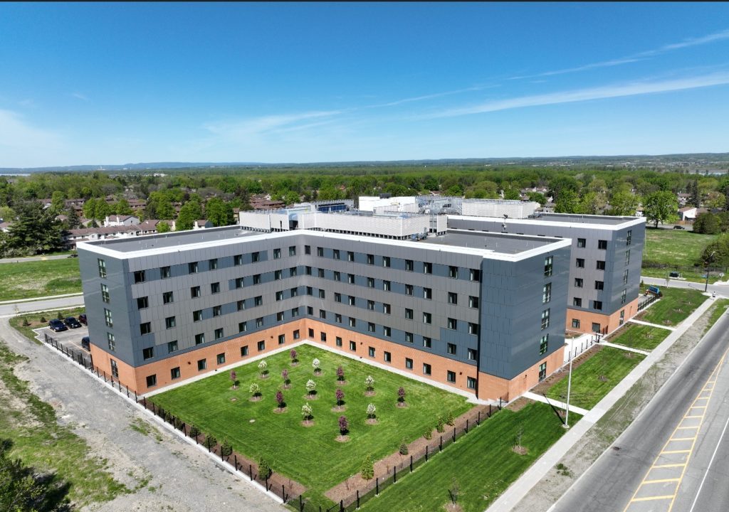 Aerial view of a modern, five-story building with gray siding, brick base, and landscaped courtyard—designed as a building for the future—situated next to a road and surrounded by greenery.