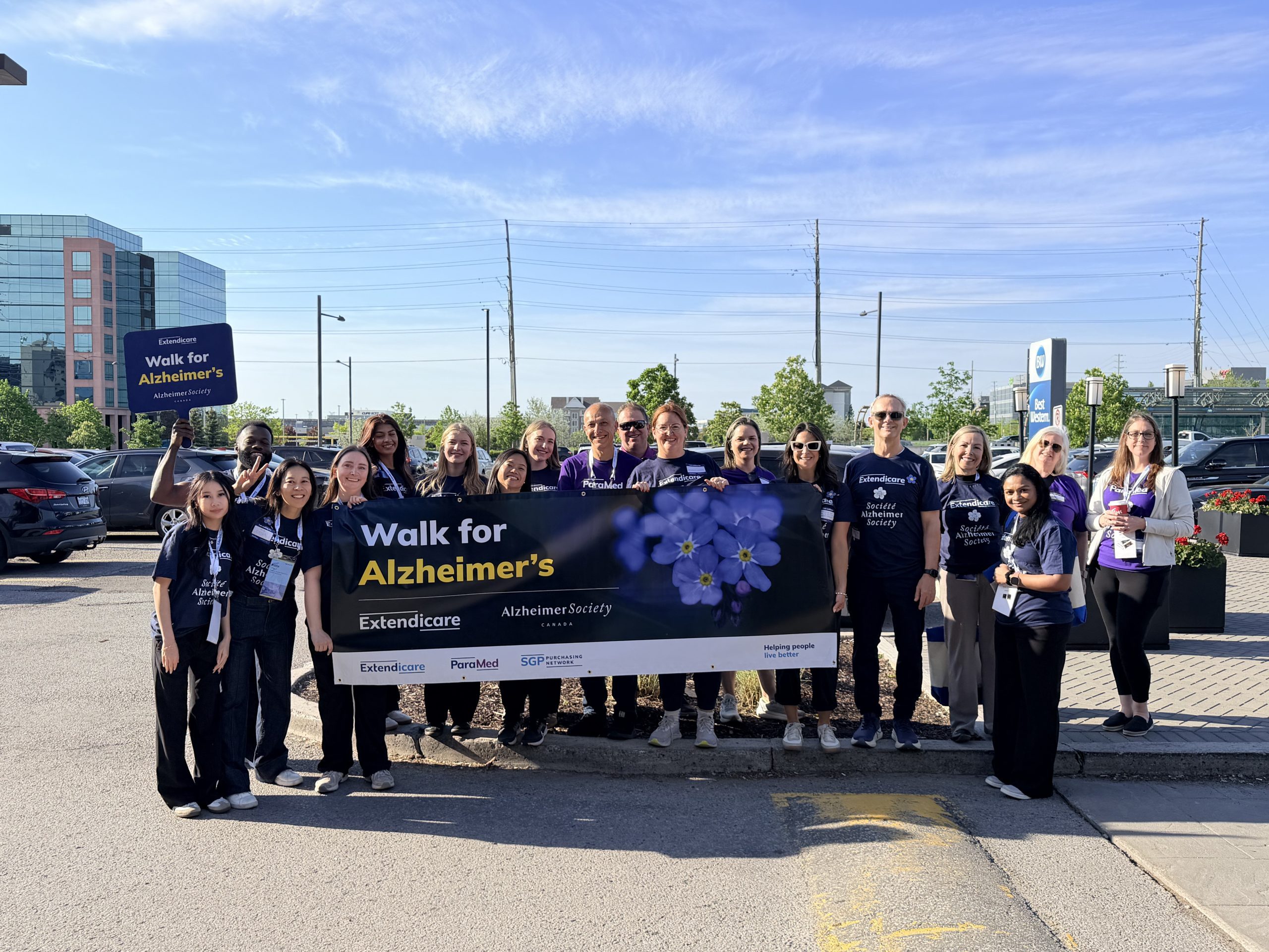 A group of people stand outdoors holding a "Walk for Alzheimer’s" banner, posing for a group photo before the event on a sunny day.